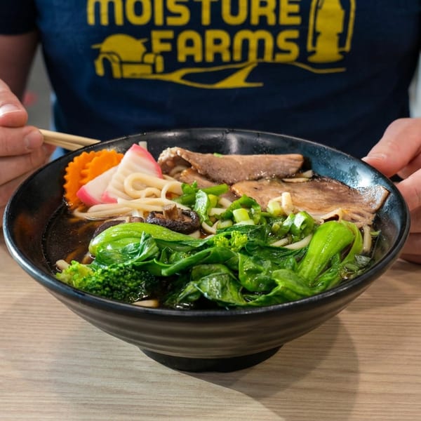 Person using chopsticks to eat a bowl of ramen with sliced meat, noodles, broccoli, greens, and vegetables at a table.