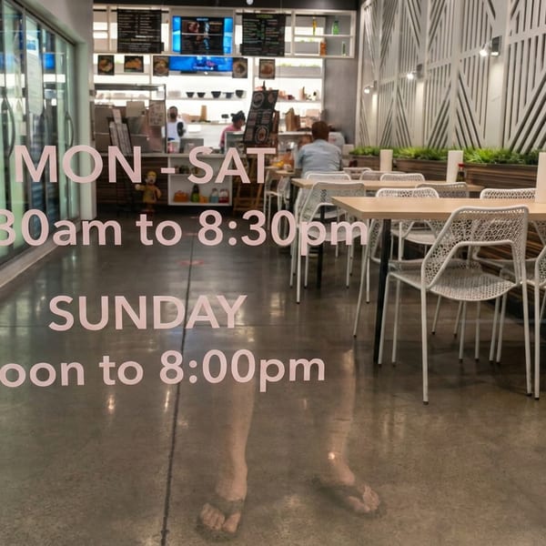 Modern restaurant dining area with white chairs, wooden tables, geometric wall decor, counter service, and hours displayed on glass.