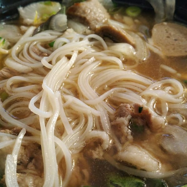 Bowl of steaming noodle soup with meat, vegetables, herbs, and chopsticks lifting noodles.