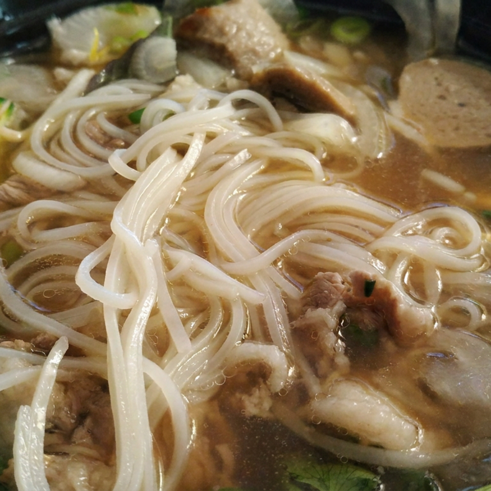 Bowl of steaming noodle soup with meat, vegetables, herbs, and chopsticks lifting noodles.