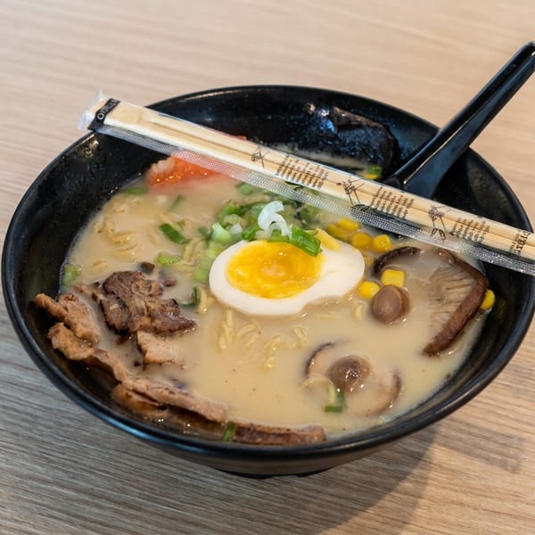 Bowl of creamy ramen topped with pork, mushrooms, corn, green onions, and a halved soft-boiled egg, with chopsticks resting across it.