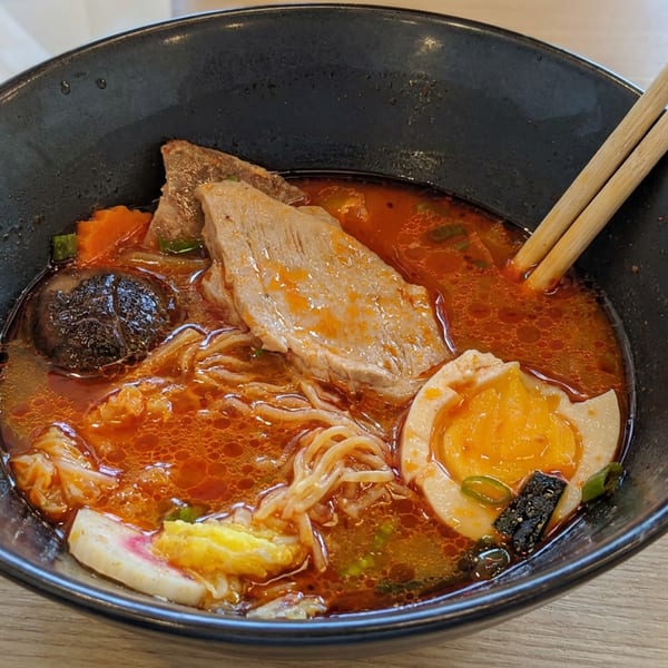 Black bowl of spicy ramen with noodles, sliced pork, egg, vegetables, and chopsticks on a wooden table.