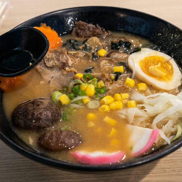 Bowl of ramen with mushrooms, corn, vegetables, sliced egg, and black spoon on a wooden table.