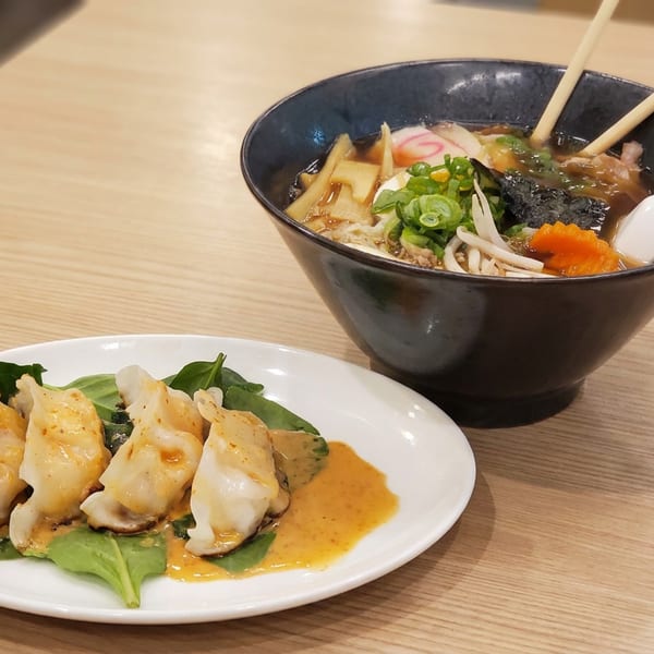 Steaming ramen bowl with chopsticks and spoon beside four dumplings on greens with orange dipping sauce.
