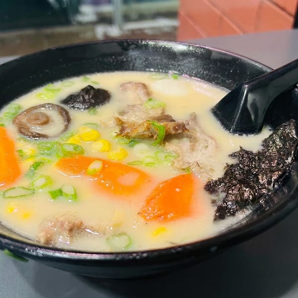 Black bowl of creamy ramen topped with carrot slices, mushrooms, corn, scallions, meat, seaweed, and a black spoon.