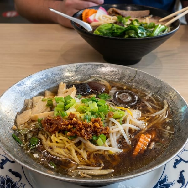 Bowl of noodle soup topped with vegetables, mushrooms, bean sprouts, scallions, and minced meat, with chopsticks and another bowl behind.