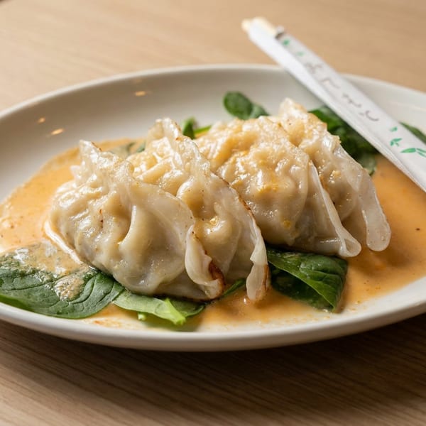 Steamed dumplings in creamy orange sauce, served over leafy greens on a white plate with wrapped chopsticks.